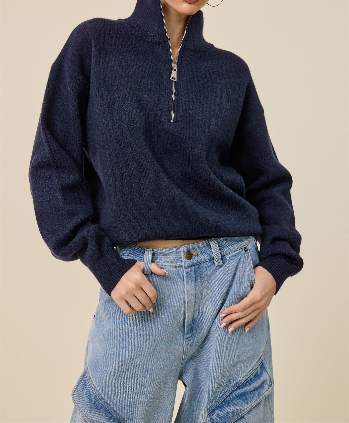 A woman modeling a navy blue quarter-zip knit sweater with a mock neck, paired with light-wash blue jeans, against a solid beige background.