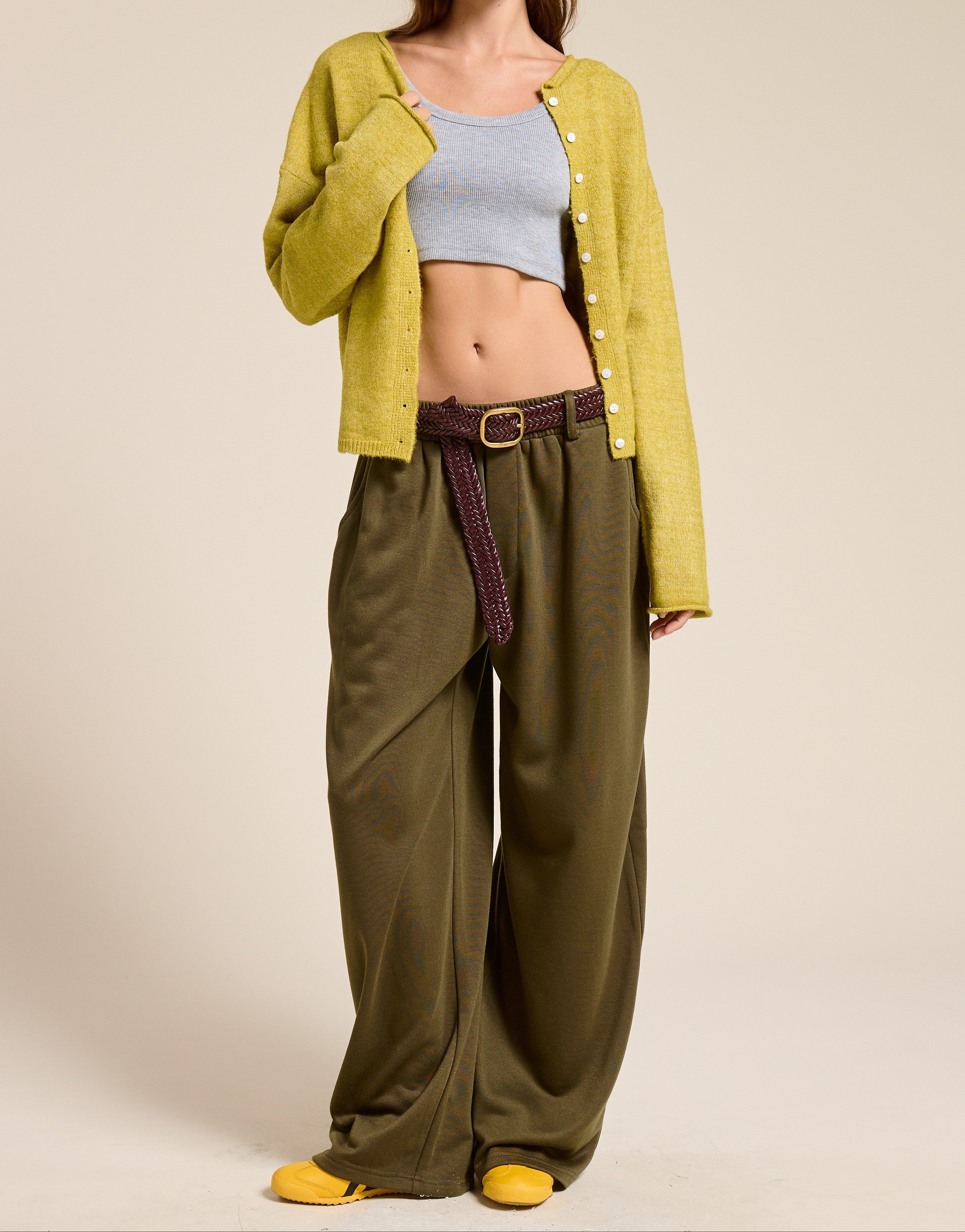 A model wearing a yellow-green cardigan, olive green wide-leg pants, a light blue crop top, a brown cap, and yellow shoes, standing against a solid beige background.