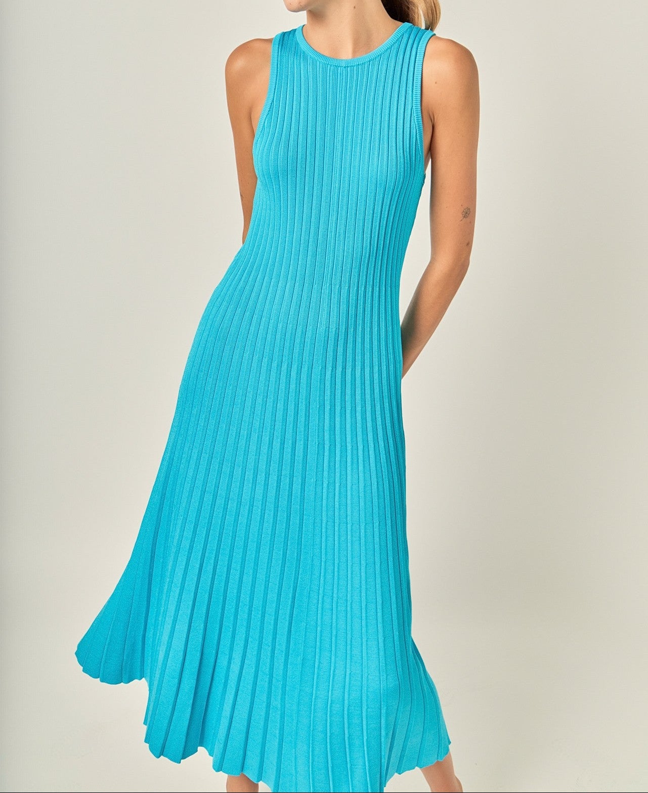 A woman wearing a sleeveless, bright turquoise blue pleated midi dress, standing against a neutral light beige studio background.