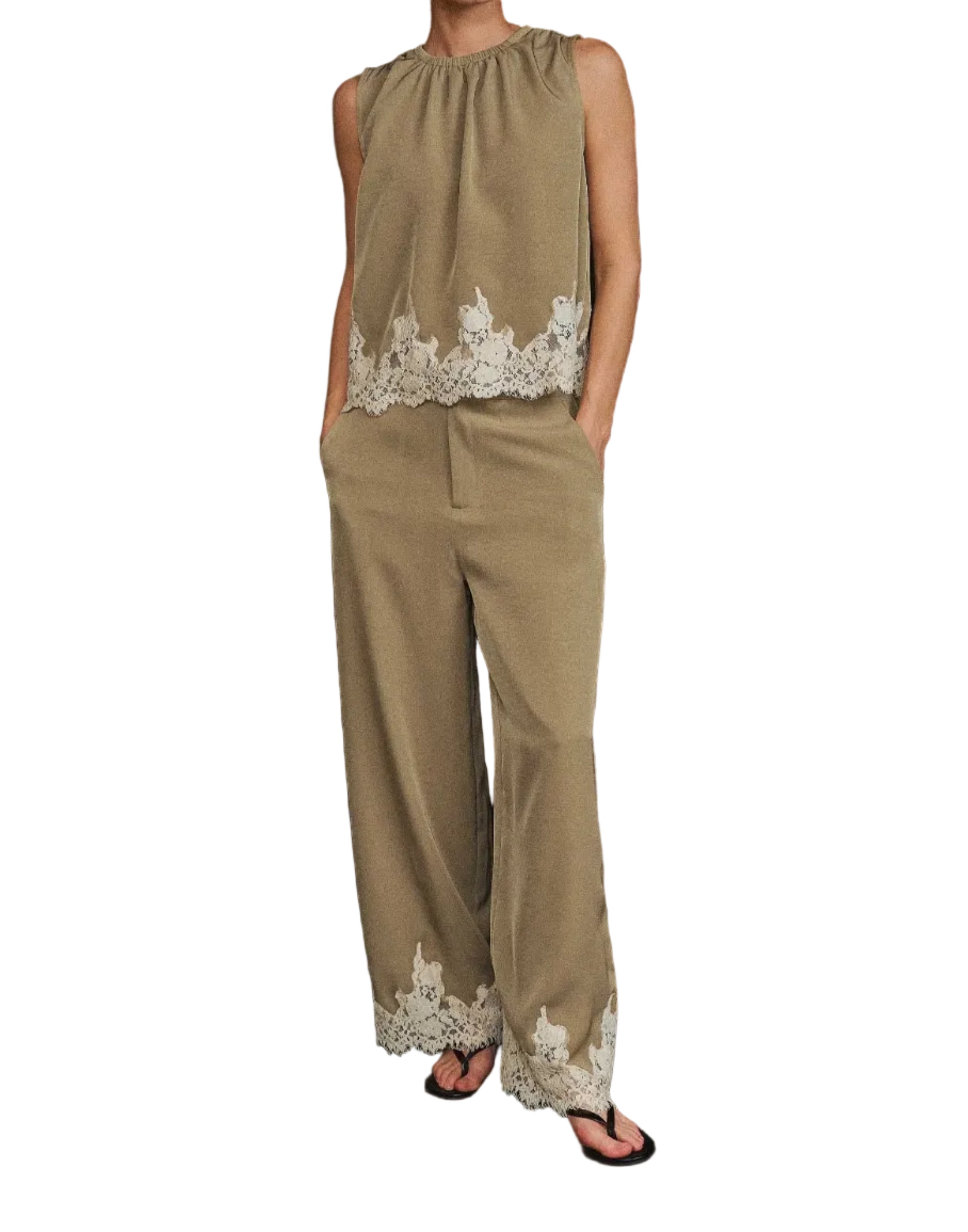 A woman modeling a sleeveless beige jumpsuit with white floral lace trim at the hem and waist, standing against a black studio backdrop.