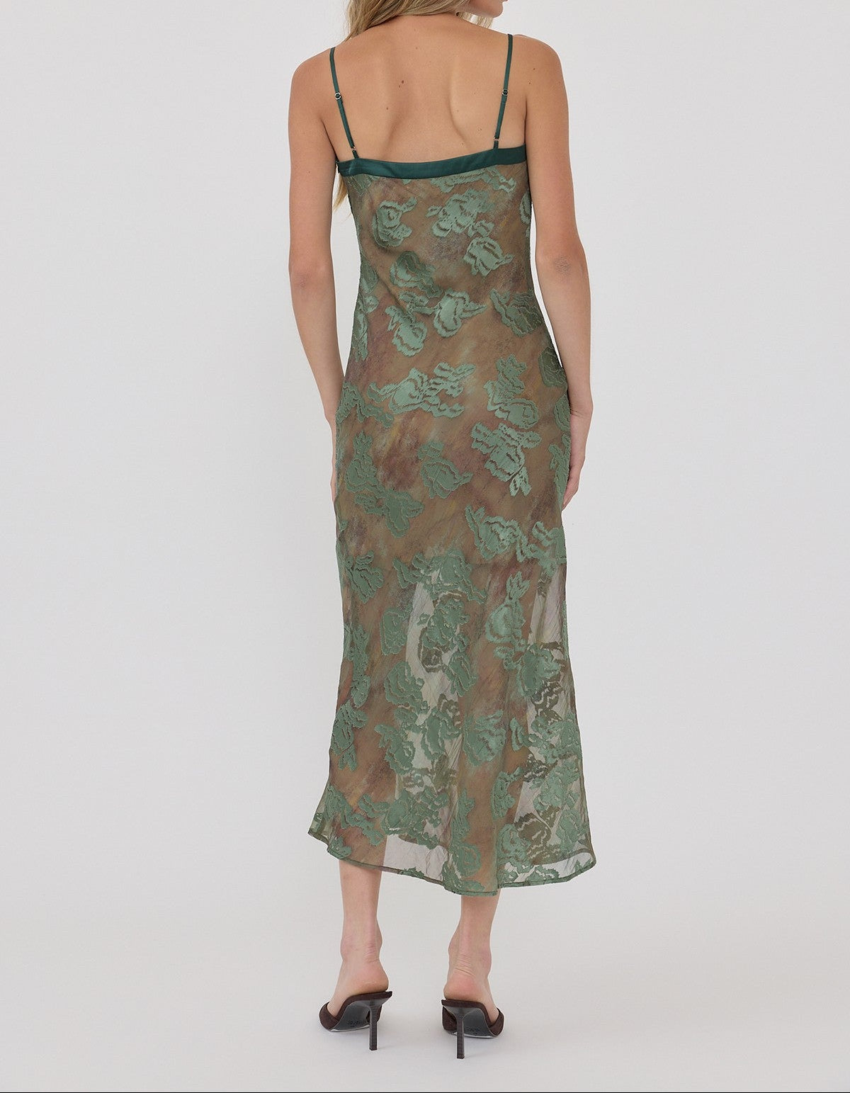 A woman viewed from the back wearing a sheer midi slip dress with a green and brown floral pattern and thin dark green straps, paired with black heels, against a light gray studio background.