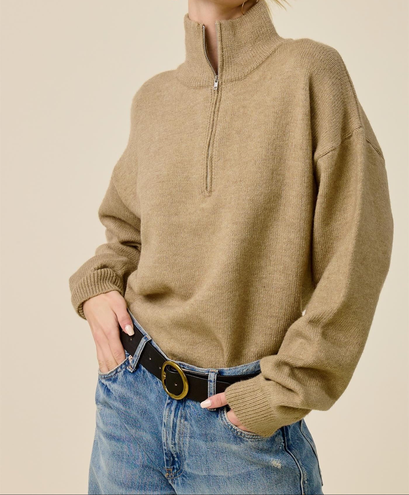 A woman wearing an oversized tan quarter-zip mock-neck sweater, blue jeans, and black sunglasses, posing against a solid beige background.