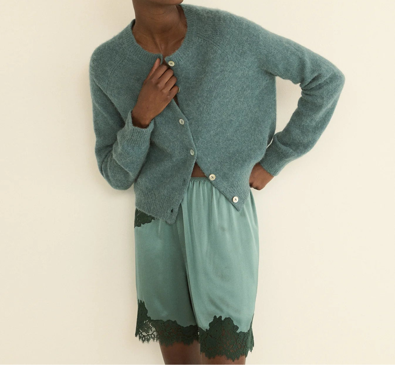 A model wearing a dusty teal knit cardigan and matching satin shorts with black lace trim, posed against a plain cream background.