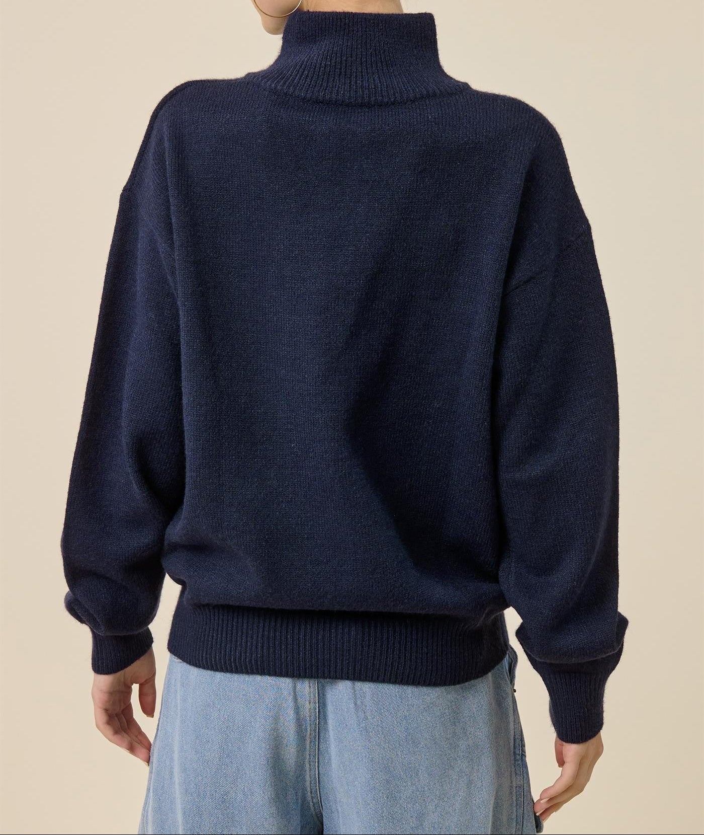 A woman shown from the back wearing a dark navy blue ribbed mock neck sweater and light blue jeans, set against a solid beige studio background.