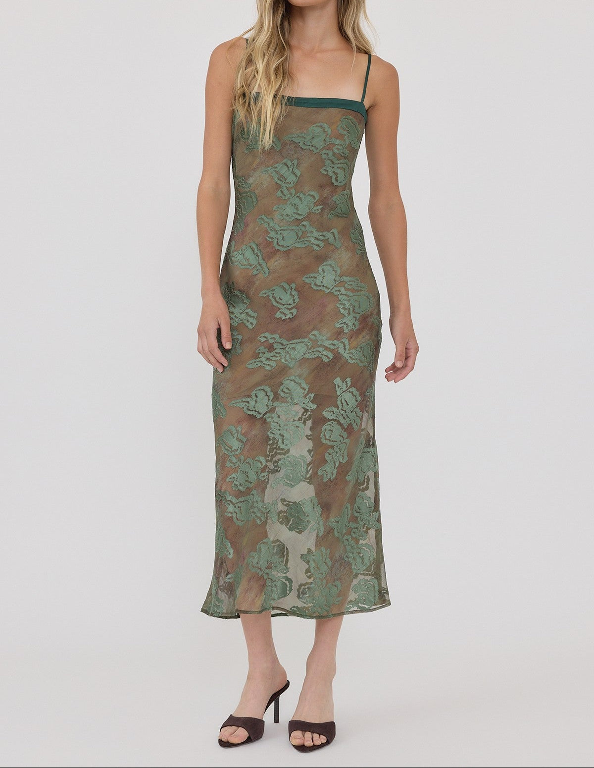 A woman modeling a sleeveless midi slip dress with a green and brown floral lace overlay, photographed against a solid light gray studio background.