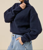 A woman wearing a navy blue knit mock-neck sweater and light blue jeans, posing against a solid beige background.