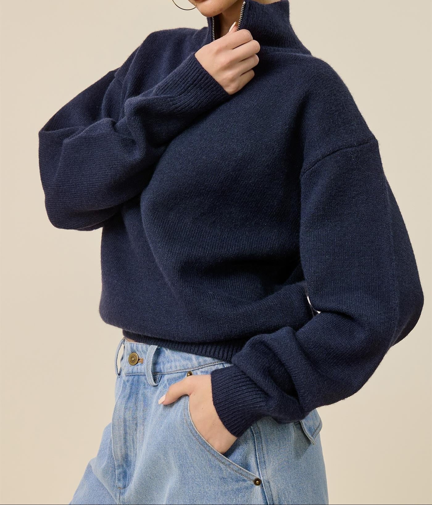 A woman wearing a navy blue knit mock-neck sweater and light blue jeans, posing against a solid beige background.