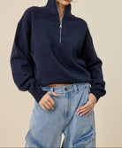 A woman modeling a navy blue quarter-zip knit sweater with a mock neck, paired with light-wash blue jeans, against a solid beige background.