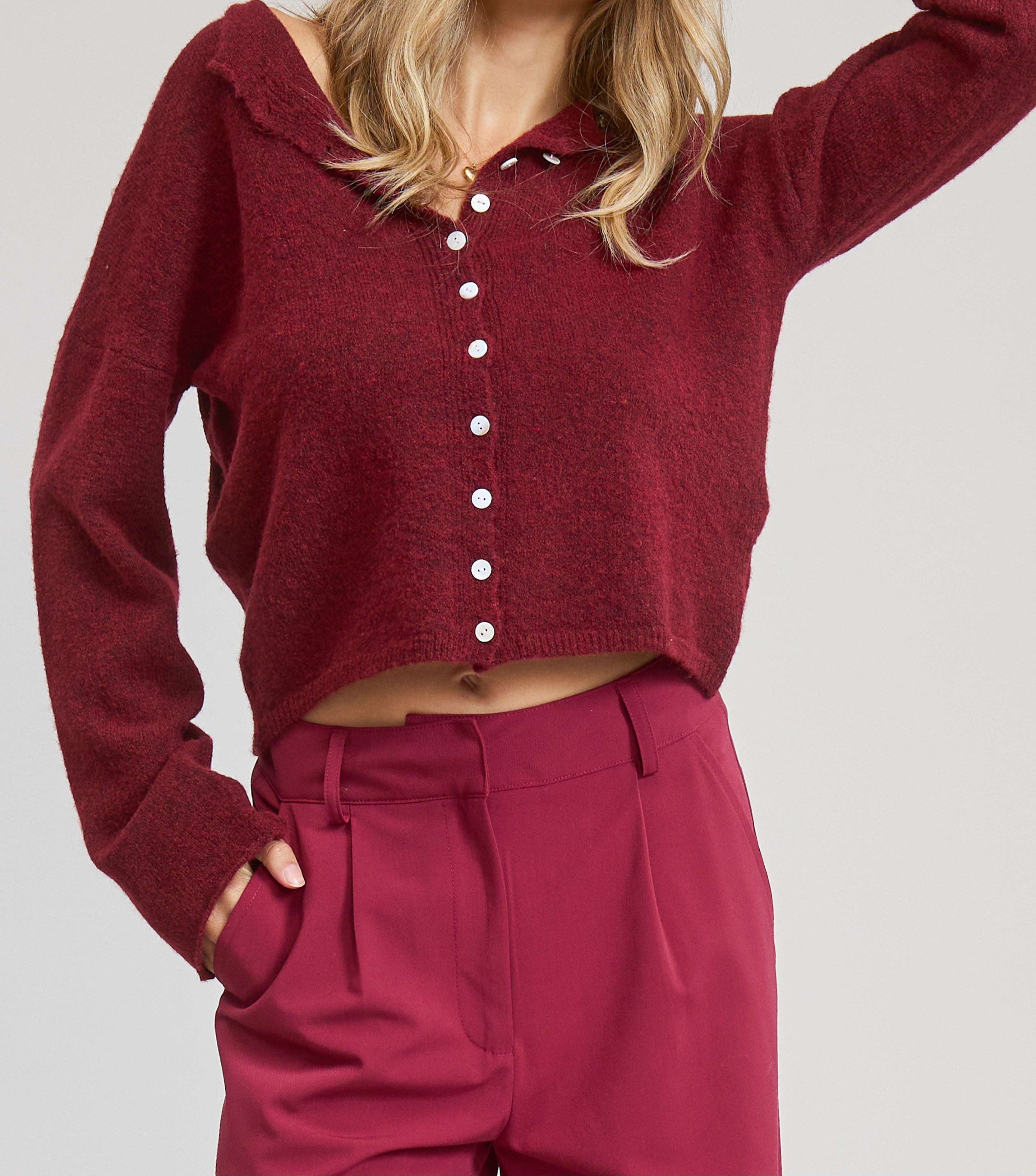 Woman wearing a burgundy cardigan and pants with a blue cap on a light gray background