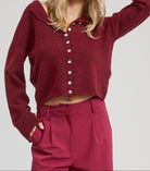 Woman wearing a burgundy cardigan and pants with a blue cap on a light gray background