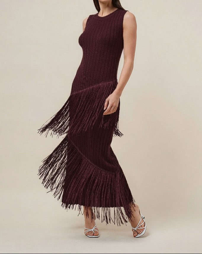 A woman modeling a sleeveless, dark burgundy maxi dress with a textured knit bodice and a long, flowing fringe skirt, set against a plain beige studio background.