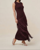 A woman modeling a sleeveless, ribbed knit maxi dress in deep burgundy with a long cascading fringe hem, paired with strappy high-heeled sandals against a solid beige studio background.