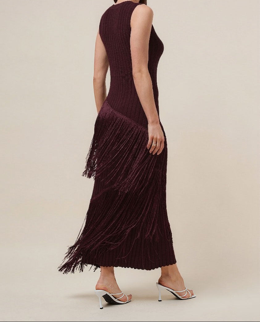 A woman modeling a sleeveless, deep burgundy maxi dress with a pleated fringe hem, shown from a three-quarters back angle against a solid beige studio background.