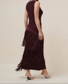 A woman modeling a sleeveless, deep burgundy maxi dress with a pleated fringe hem, shown from a three-quarters back angle against a solid beige studio background.