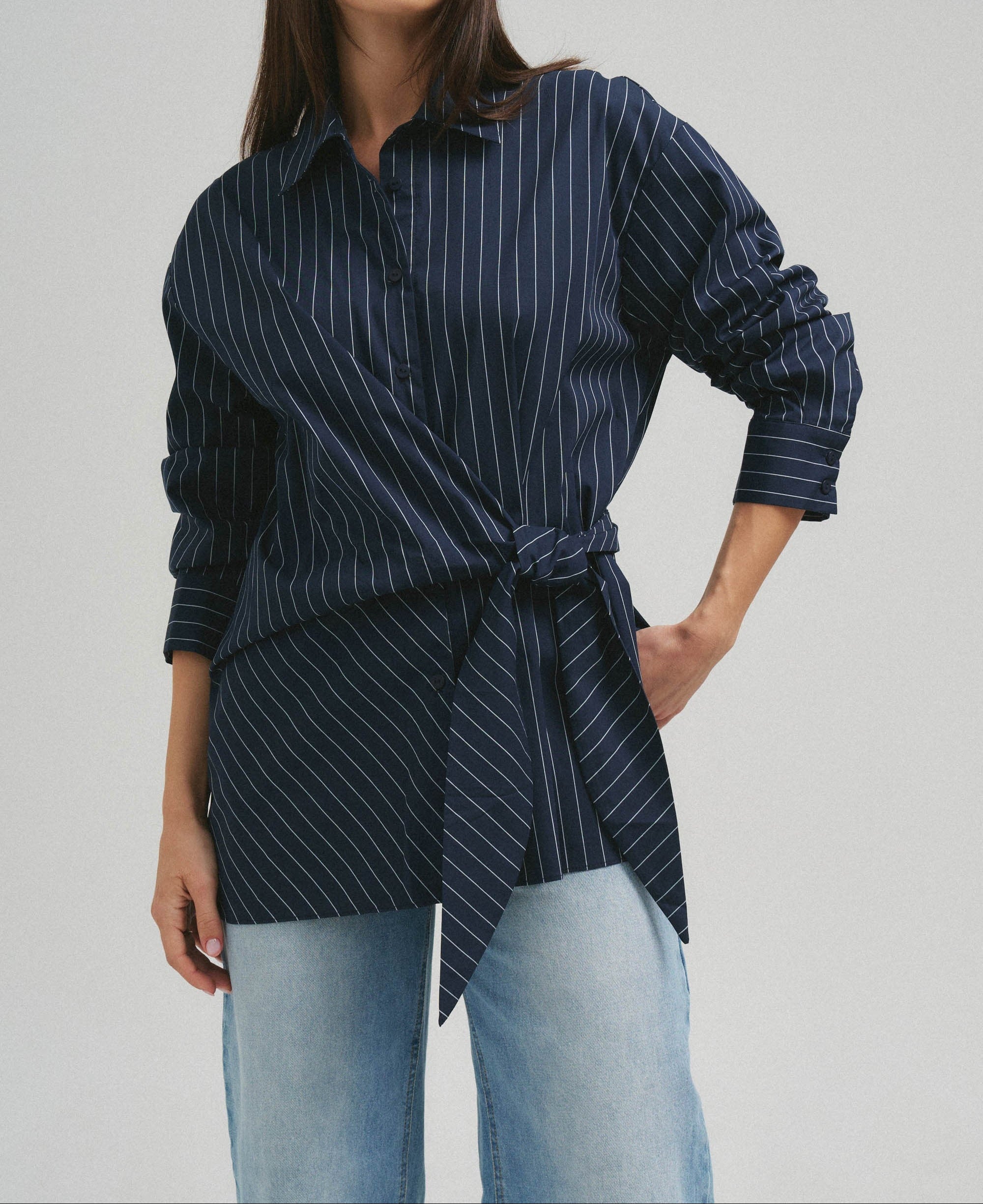 A woman modeling a navy blue long-sleeve button-up shirt with thin white vertical pinstripes, styled with light-wash blue jeans against a solid light gray studio background.