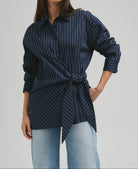 A woman modeling a navy blue long-sleeve button-up shirt with thin white vertical pinstripes, styled with light-wash blue jeans against a solid light gray studio background.