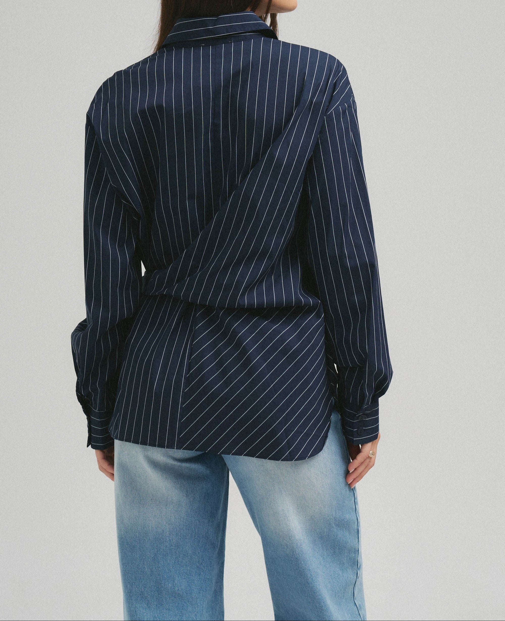 A woman shown from the back wearing a navy blue long-sleeve shirt with thin white vertical pinstripes and light blue jeans, against a plain light gray studio background.