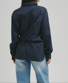 A woman shown from the back wearing a navy blue long-sleeve shirt with thin white vertical pinstripes and light blue jeans, against a plain light gray studio background.