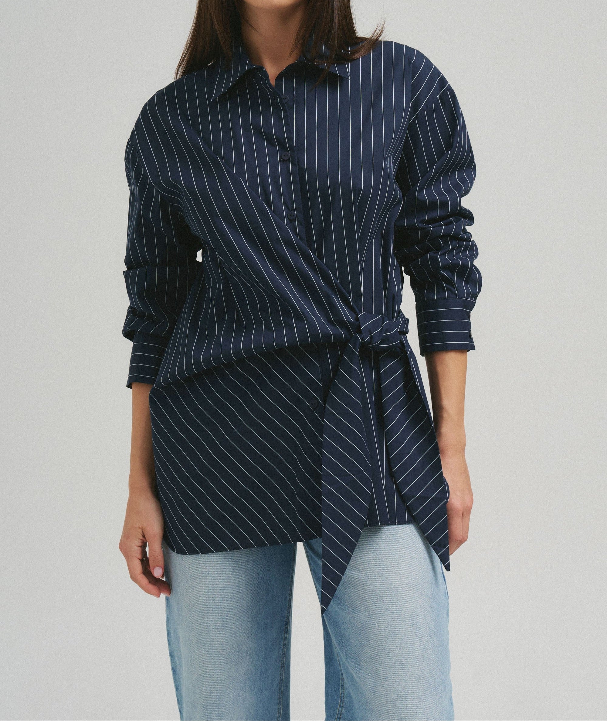 A woman wearing a navy blue and white pinstriped button-up shirt with a twisted waist detail, paired with light blue jeans, against a plain light gray studio background.
