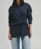 A woman wearing a navy blue and white pinstriped button-up shirt with a twisted waist detail, paired with light blue jeans, against a plain light gray studio background.