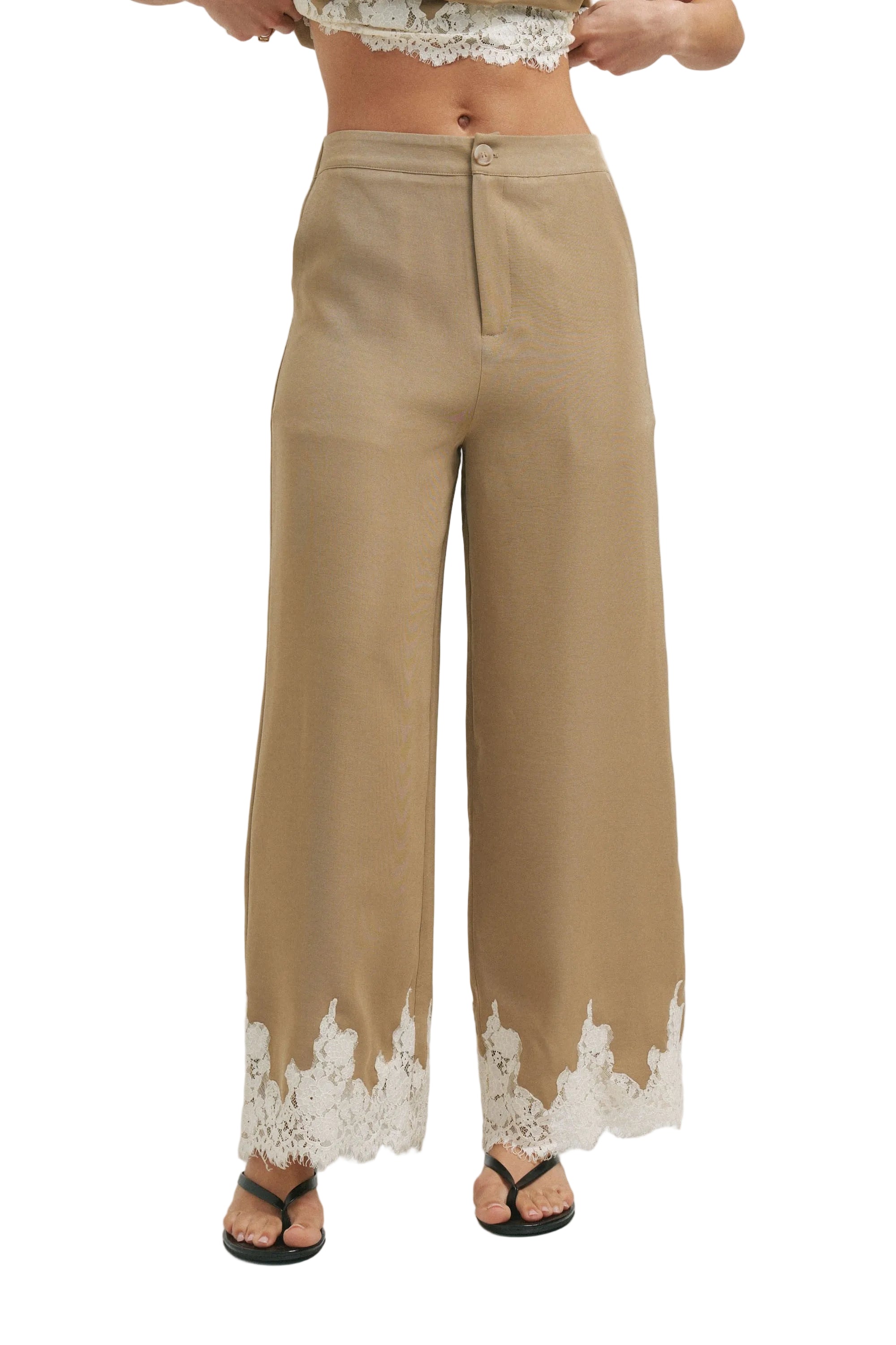 A person wearing high-waisted, wide-leg tan pants with white lace trim at the hem, paired with black flip-flop sandals against a light gray studio background.