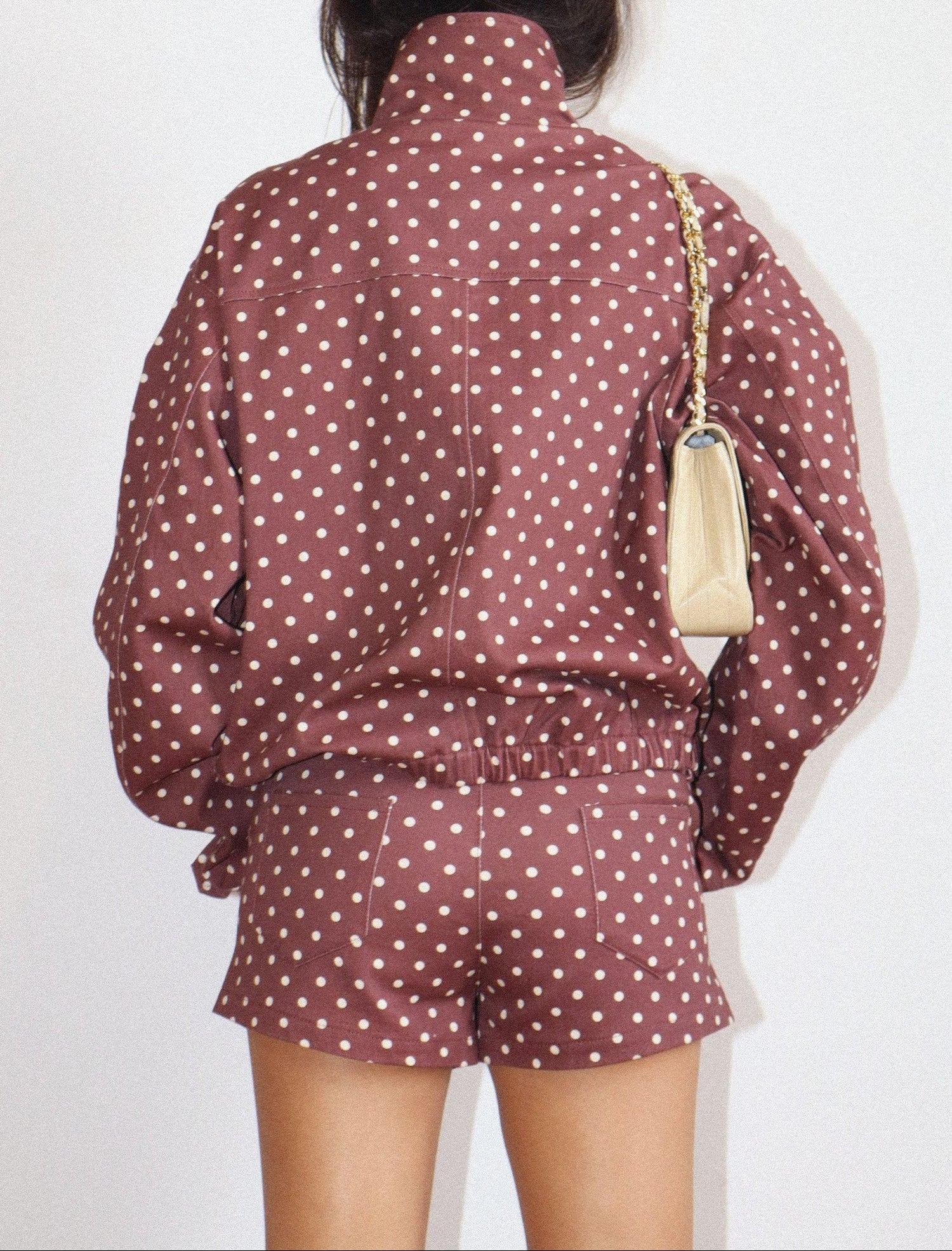 A person shown from the back wearing a matching long-sleeve collared top and shorts set in a dusty rose-brown color with white polka dots. A small beige shoulder bag is also visible against a plain off-white background.