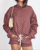 A woman wearing a matching dusty rose and white polka dot jacket and shorts set, holding a cream-colored shoulder bag against a plain white background.