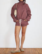 A woman stands against a white wall wearing a matching long-sleeve turtleneck top and shorts set in a dusty rose color with white polka dots. She is carrying a small cream shoulder bag and wearing beige flats.