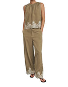A woman modeling a sleeveless beige jumpsuit with white floral lace trim at the hem and waist, standing against a black studio backdrop.