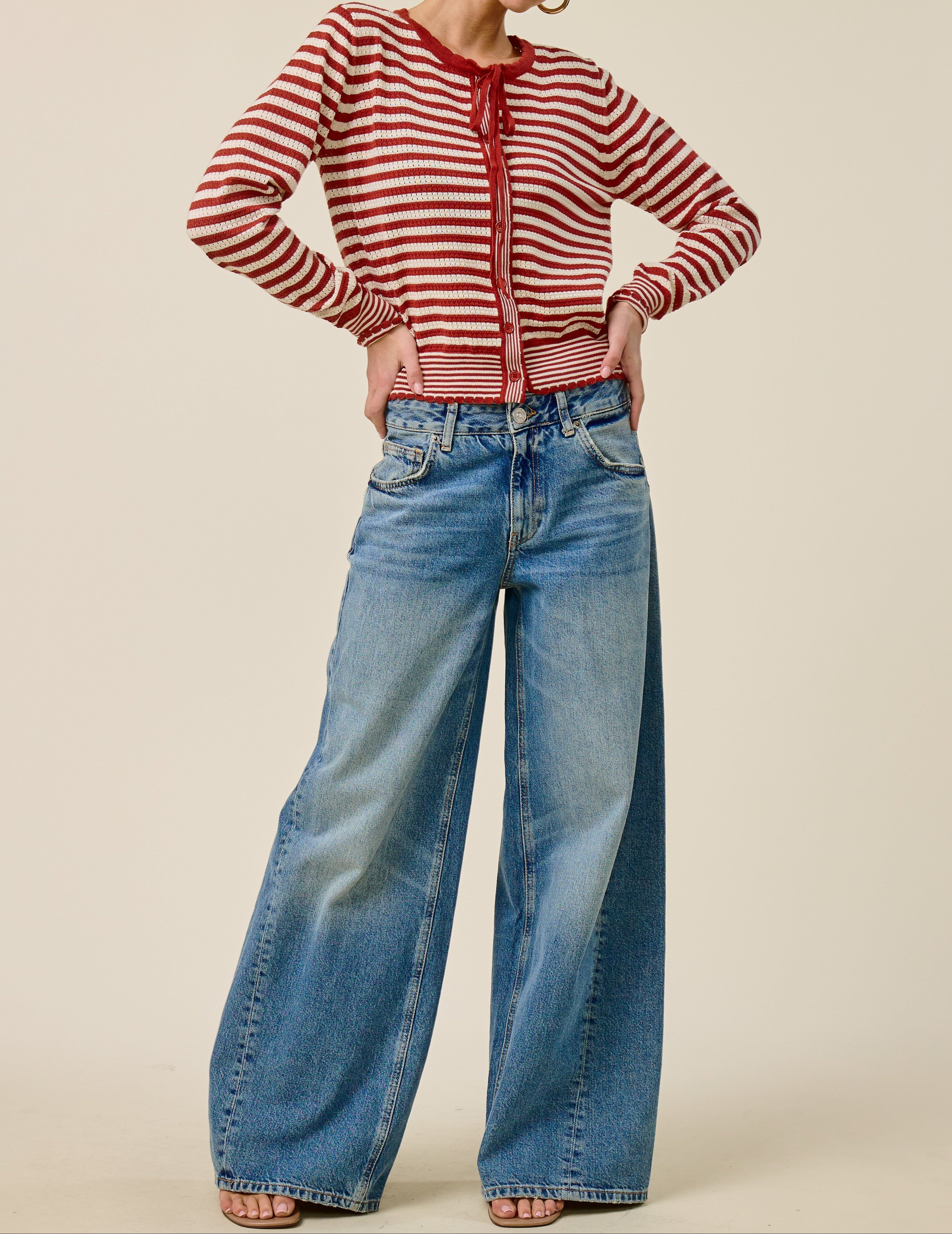 A model wearing a red and white striped knit cardigan with a tie-neck detail, paired with high-waisted, wide-leg blue jeans, against a solid beige studio background.