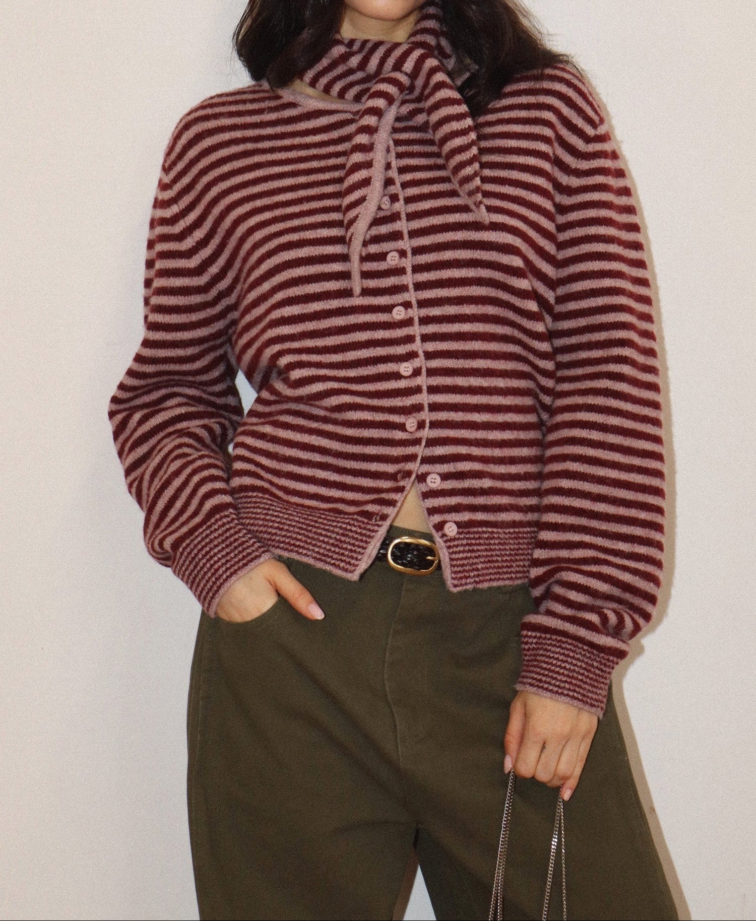 A woman wearing a cropped, long-sleeve knit cardigan with horizontal burgundy and beige stripes, featuring a high ribbed mock neck with a tie detail and front buttons. She is also wearing olive green trousers.