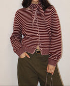 A woman wearing a cropped, long-sleeve knit cardigan with horizontal burgundy and beige stripes, featuring a high ribbed mock neck with a tie detail and front buttons. She is also wearing olive green trousers.