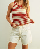 A woman modeling a red and white striped ribbed tank top with a small American flag graphic on the chest, paired with white denim shorts, against a light gray studio background.