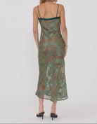 A woman viewed from the back wearing a sheer midi slip dress with a green and brown floral pattern and thin dark green straps, paired with black heels, against a light gray studio background.