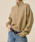 A woman wearing an oversized tan quarter-zip mock-neck sweater, blue jeans, and black sunglasses, posing against a solid beige background.