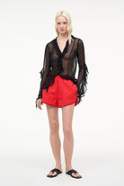 Woman wearing a black sheer top with lace details and red shorts on a white background