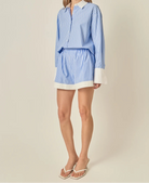 A woman modeling a light blue and white vertically striped shirt dress with a white collar, white cuffs, and a white hem, paired with white heeled sandals, against a solid beige background.