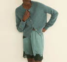 A model wearing a dusty teal knit cardigan and matching satin shorts with black lace trim, posed against a plain cream background.