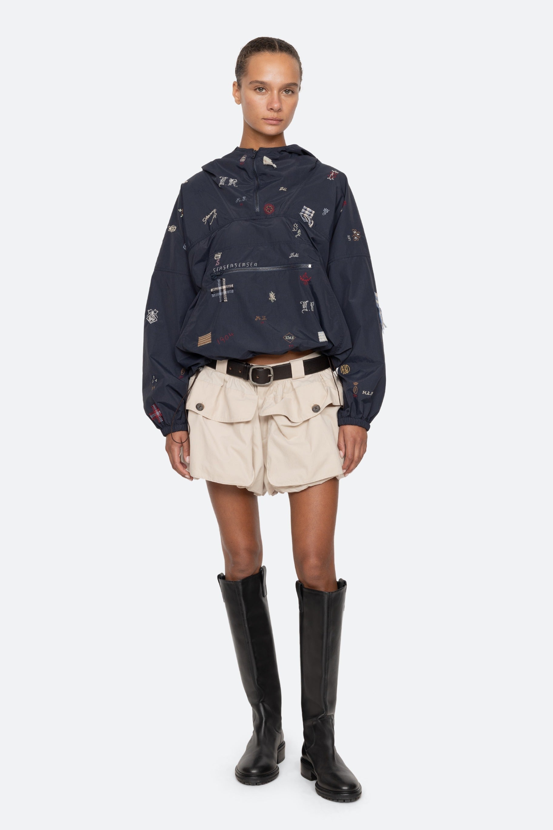 Person wearing a dark blue patterned jacket, beige shorts, and black boots on a white background