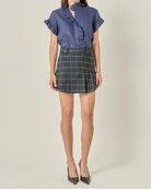 A model stands against a light beige studio background wearing a blue short-sleeve blouse with ruffle details and a dark green plaid mini skirt, paired with dark pointed-toe heels.