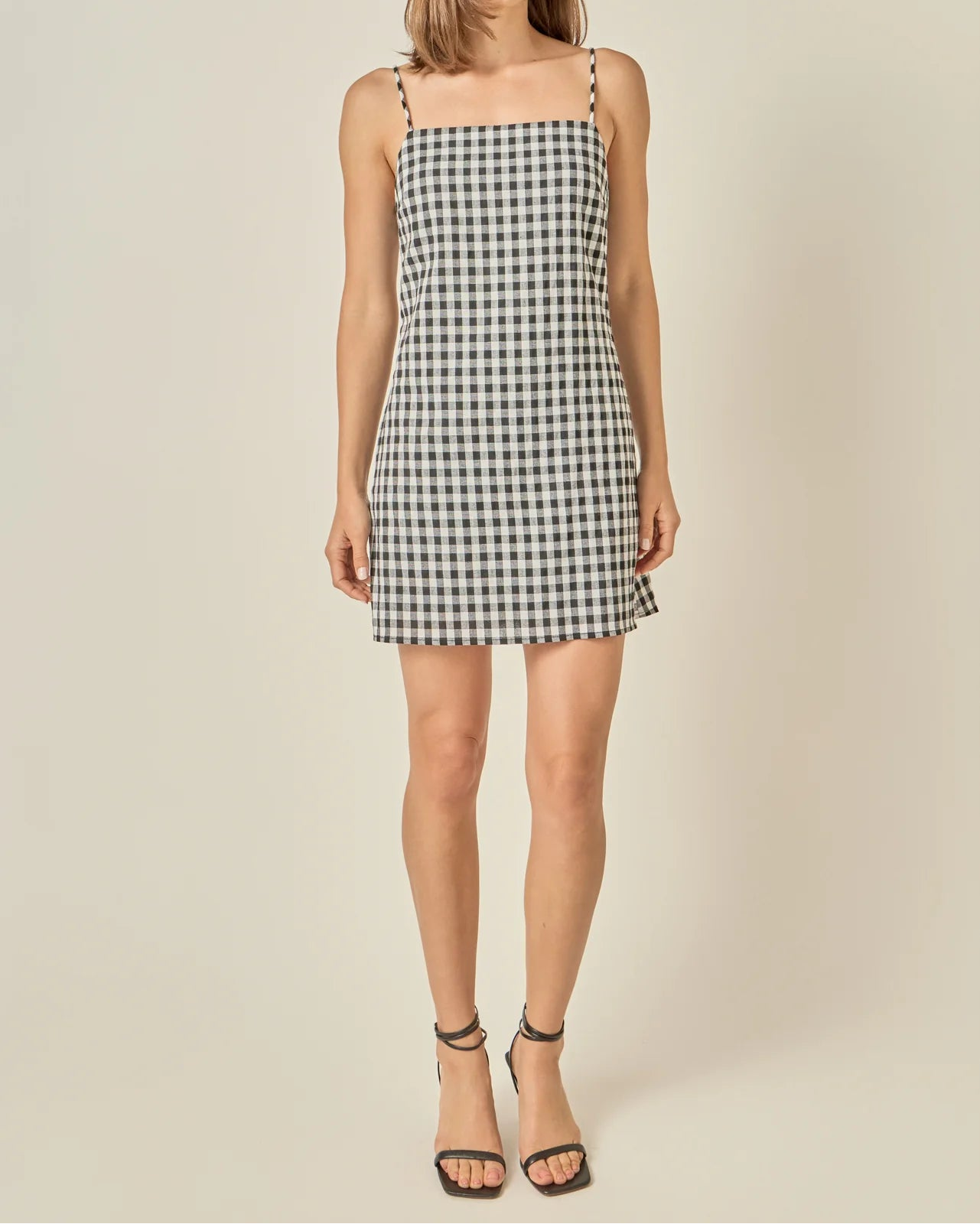 A woman modeling a sleeveless black and white gingham mini dress with thin shoulder straps, paired with black strappy high-heeled sandals, against a solid light beige studio background.