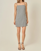 A woman modeling a sleeveless black and white gingham mini dress with thin shoulder straps, paired with black strappy high-heeled sandals, against a solid light beige studio background.