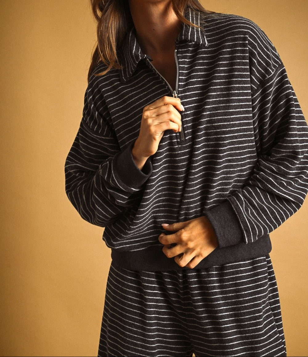 A woman wearing a dark charcoal gray and white striped zip-up sweatshirt and matching striped sweatpants, paired with a maroon baseball cap, against a solid warm brown studio background.