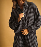 A woman wearing a dark charcoal gray and white striped zip-up sweatshirt and matching striped sweatpants, paired with a maroon baseball cap, against a solid warm brown studio background.