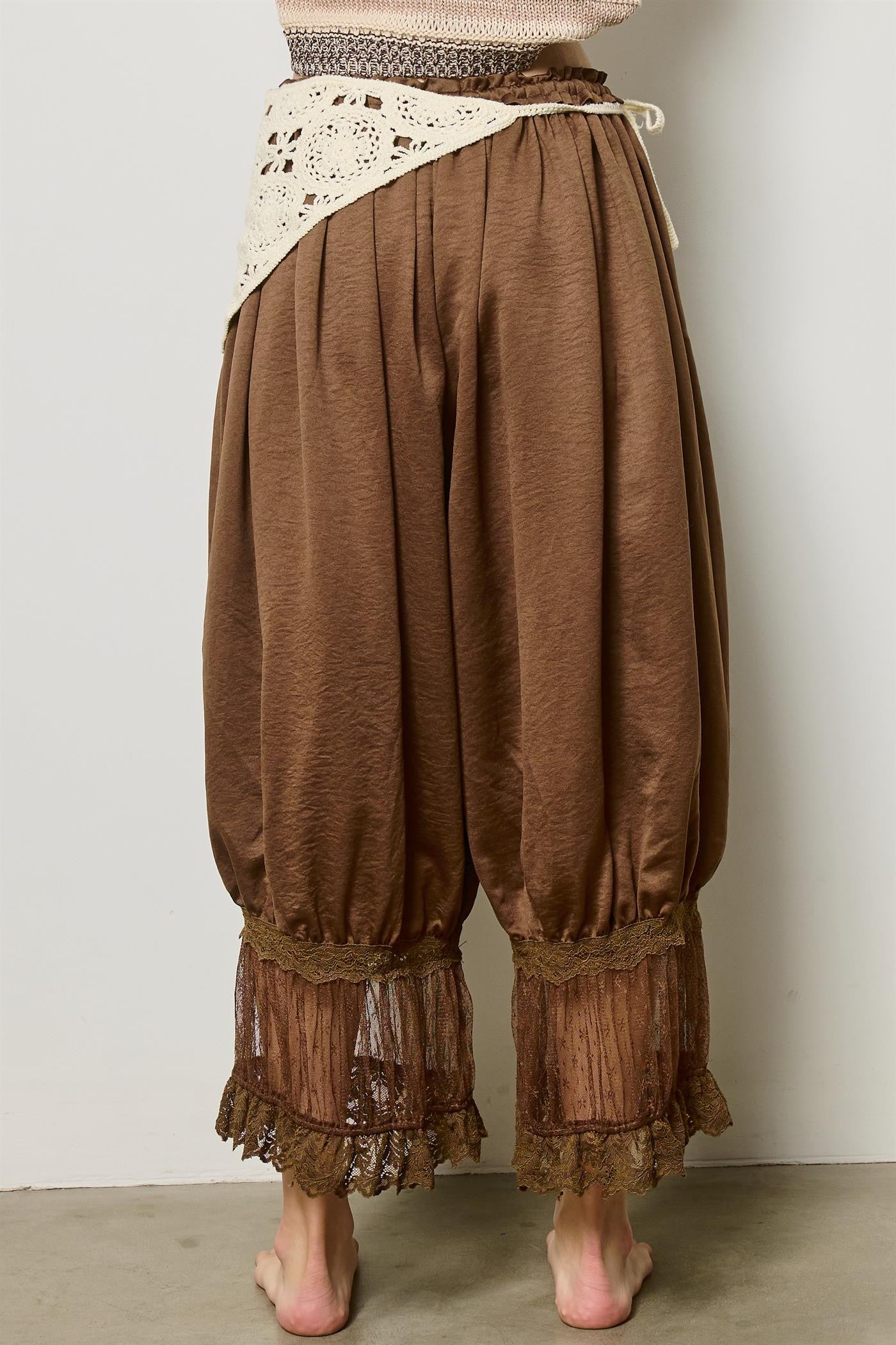 Brown harem-style pants with a cream lace waist overlay and sheer lace cuffs, shown on a model from the back against a plain light gray wall.