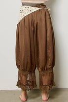 Brown harem-style pants with a cream lace waist overlay and sheer lace cuffs, shown on a model from the back against a plain light gray wall.