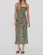A woman modeling a sleeveless midi slip dress with a green and brown floral lace overlay, photographed against a solid light gray studio background.