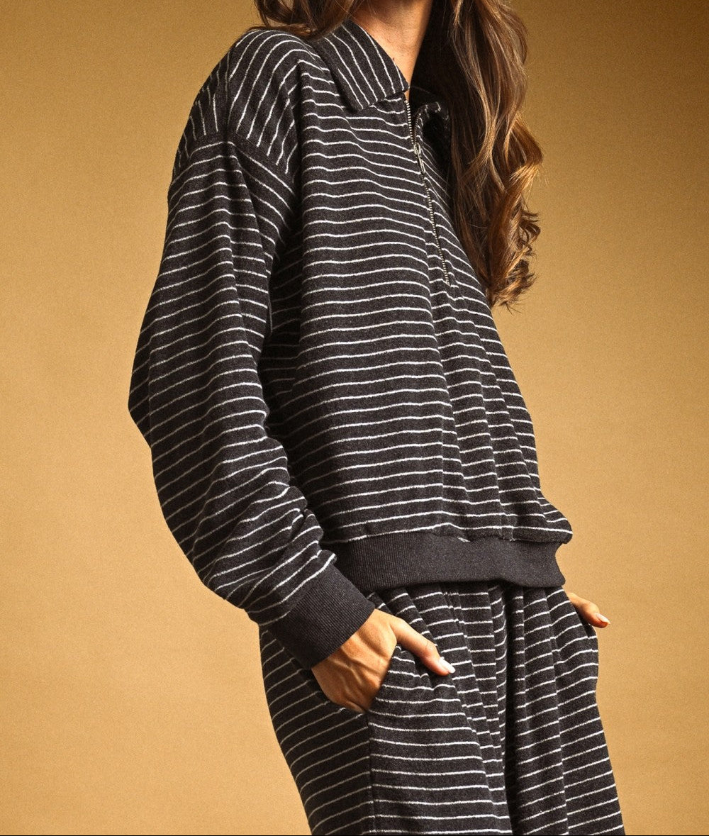 A woman wearing a dark gray and white striped lounge set, featuring a long-sleeve half-zip sweatshirt and matching sweatpants, paired with a maroon baseball cap, set against a solid warm brown background.