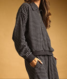 A woman wearing a dark gray and white striped lounge set, featuring a long-sleeve half-zip sweatshirt and matching sweatpants, paired with a maroon baseball cap, set against a solid warm brown background.
