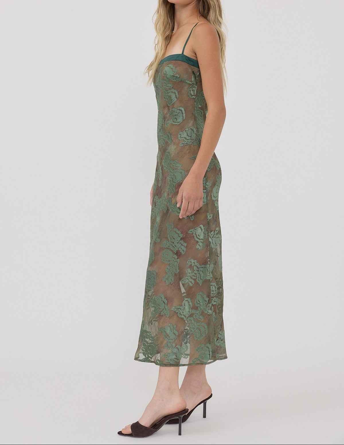 A woman modeling a sheer green midi slip dress with a tonal floral pattern, paired with dark brown mule heels, against a plain light gray studio background.